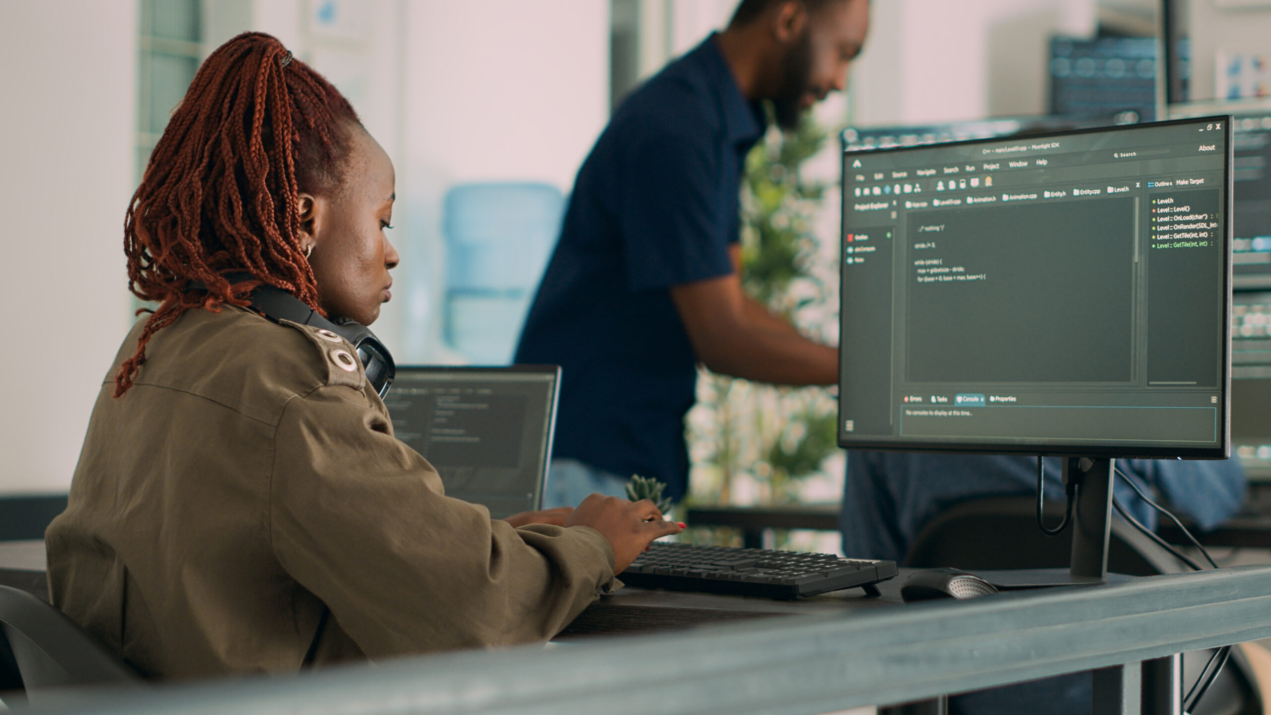 Imagem capa do conteúdo sobre Operações de segurança, onde há uma mulher negra de tranças vermelhas vestindo um casaco bege está sentada em frente ao computador, na tela aparecem códigos que ela está escrevendo no teclado.