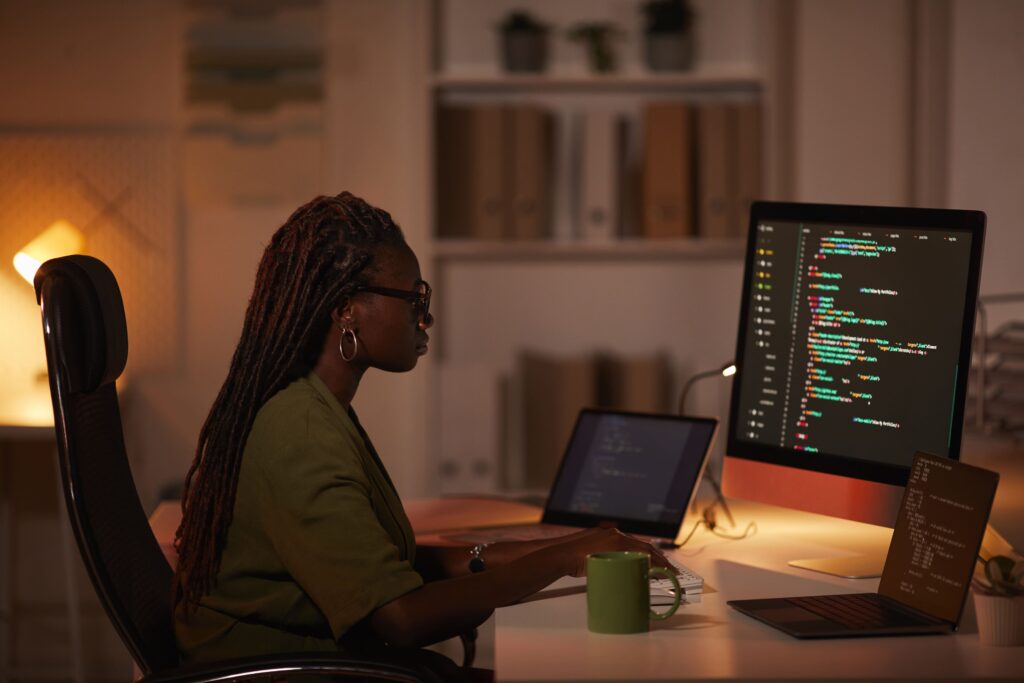 Imagem de capa do artigo sobre como fazer testes automatizados de API Rest com uma ferramenta de IA, a StackSpot AI. Vista lateral de uma mulher afro-americana contemporânea escrevendo código e olhando para a tela do computador enquanto trabalhava em um escritório escuro.