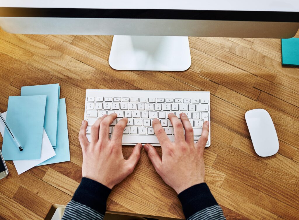 Capa do artigo “Práticas de SRE na modernização de legados com a StackSpot Cloud Services” onde vemos uma pessoa de pele branca trabalhando em uma mesa de escritório. Na mesa temos um monitor, um teclado, blocos de anotações coloridos e um mouse.