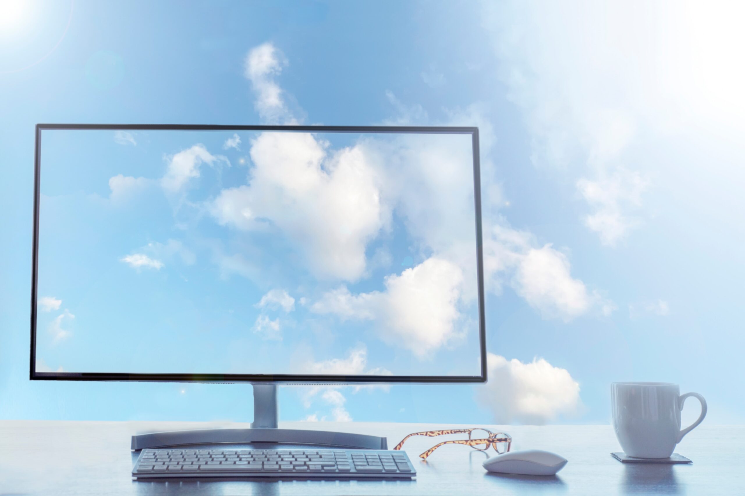 Capa do artigo “Migração para a nuvem: como implementar um plano de sucesso?” onde vemos um monitor de computador transparente em cima de uma mesa com teclado, mouse e um par de óculos. No fundo da imagem vemos um céu com nuvens, exibido também no monitor.
