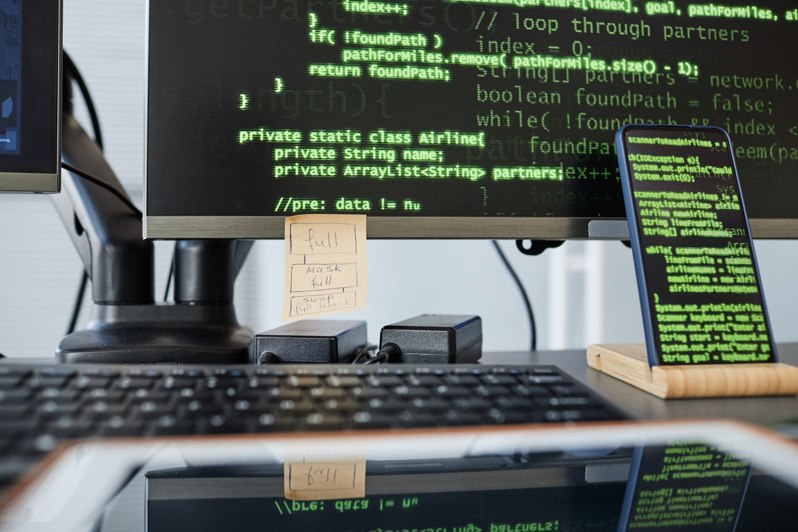 Imagem capa do conteúdo sobre Foto de uma tela de computador e de um celular com códigos em verde. No monitor do computador, há um post-it e abaixo o teclado.
