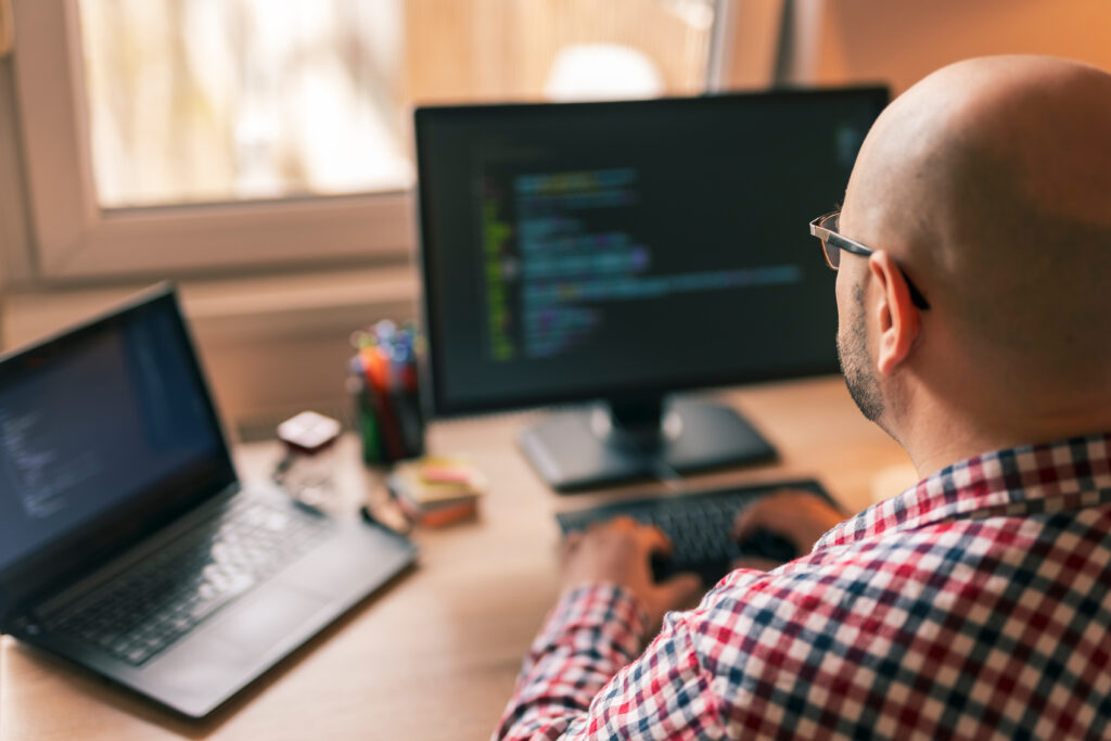 Capa do artigo "Modernização de aplicações com IA: 5 vantagens para as empresas". Na imagem, aparece um homem branco e calvo de camisa xadrez em frente ao monitor com códigos e com as mãos no teclado.