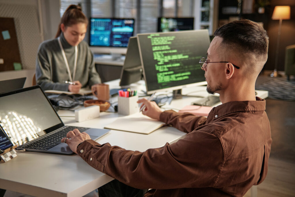 Cover of the article “How Test-Driven Development (TDD) and prompt engineering have improved software development”. The photo shows a desk with a white man with short brown hair in the foreground, he is in front of a laptop. In the background is a woman with long brown hair writing in a notebook. In the background we see computer monitors.