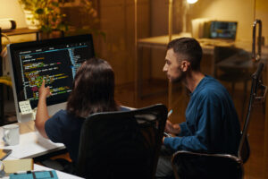 Cover of the developer platform article with a man and a woman, both white, sitting in office chairs in front of a monitor displaying programming codes. The woman points at the screen and the man watches.