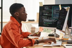 Cover of the article on How to use AI in documentation. The image shows a young black man wearing glasses and an orange shirt. He is standing in front of computer monitors in an office environment.