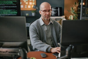 Imagem capa do conteúdo sobre Automatização de Documentação com Gen AI, onde há um homem branco, de óculos, vestindo um suéter de frente para um computador enquanto trabalho com desenvolvimento de software.
