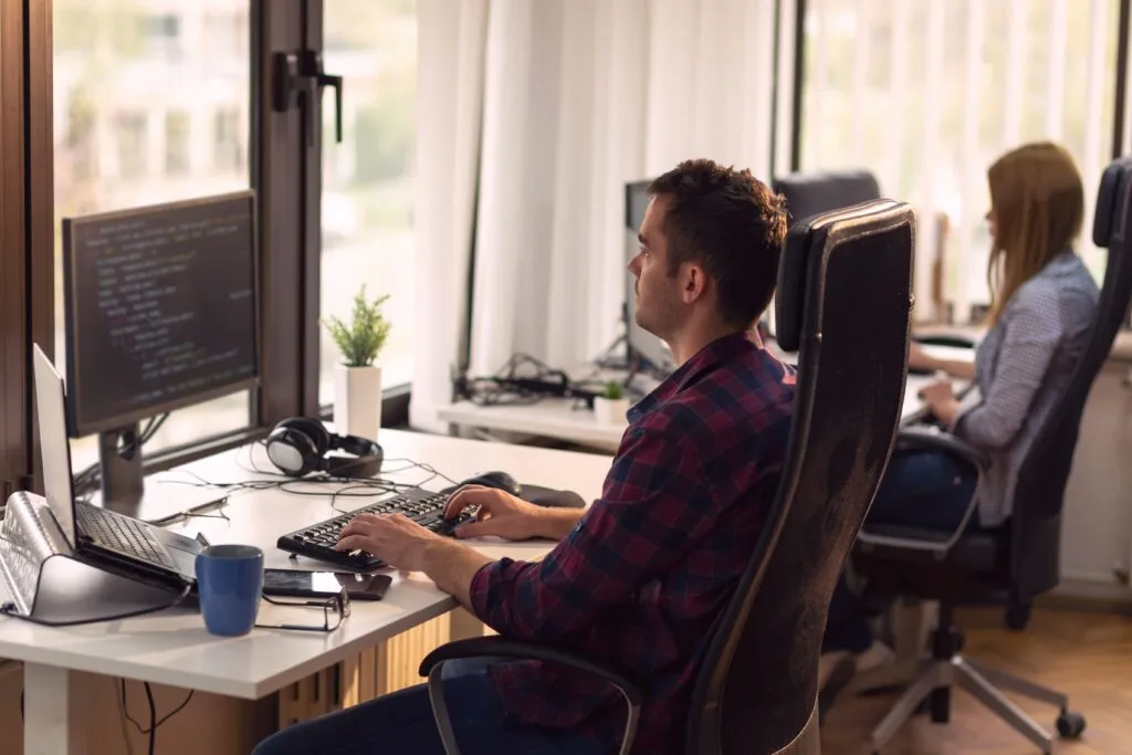 Cover image of the content about "Platform Engineering Team" featuring a white man seated in front of a computer screen with code. To his right, further in the background of the image, a woman is seated facing a monitor.