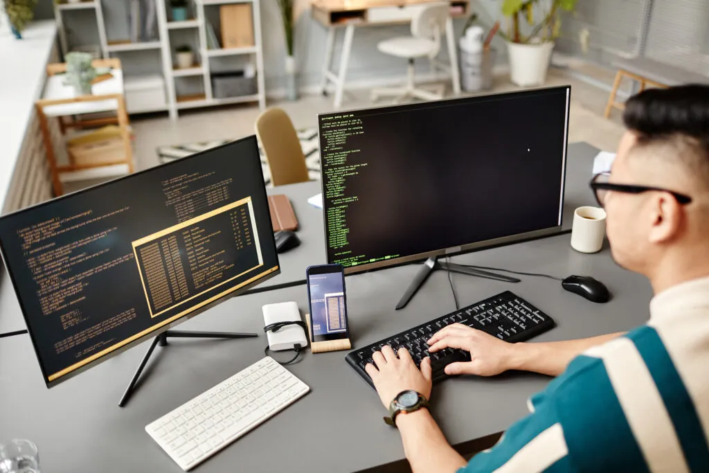 Capa do artigo Vantagens da plataforma de desenvolvimento com uma foto de uma pessoa de características asiáticas em frente ao computador, ela possui dois monitores e dois teclados em cima da mesa. As mãos da pessoa está em um dos teclados.