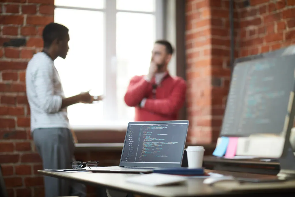 Cover image for the content 'Key traits of successful CIOs in Legacy Modernization projects,' featuring two men conversing in the background. Prominently displayed is an open notebook with code.