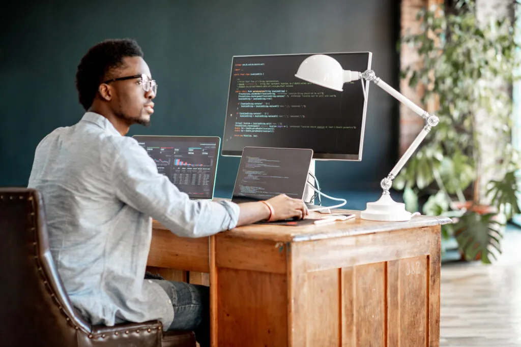 Cover of the article on CTOs and Generative AI: 3 misconceptions to overcome and 5 reasons to embrace AI now. The image shows a black man with short hair and wearing glasses in front of an office desk with monitors, a notebook and a lamp. The man looks away and has his arm on the table.