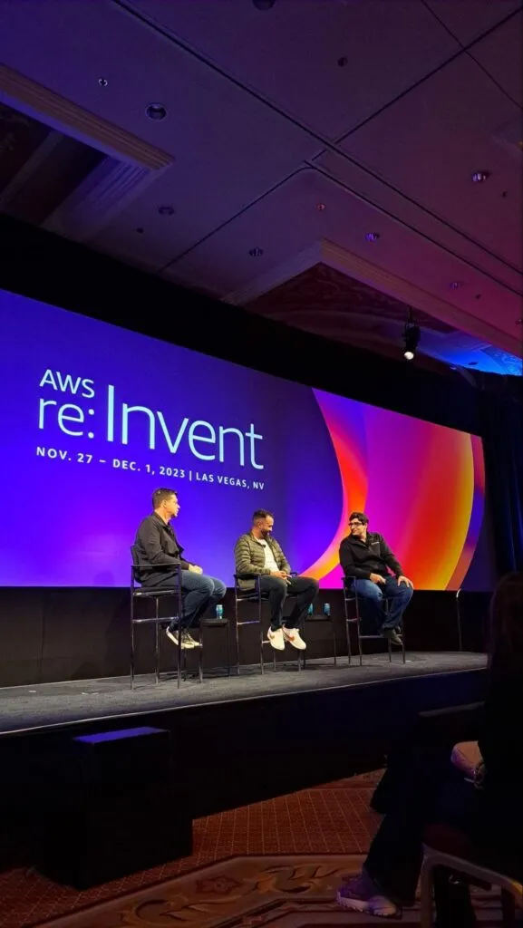 Photo of StackSpot's press conference at AWS re:Invent 2023. Three men are sitting on chairs in front of an audience; the background shows the visual identity of the event in purple, orange, pink and red.