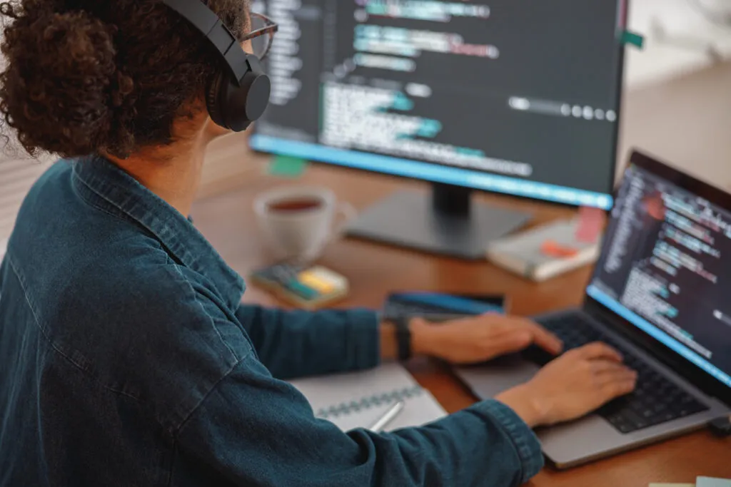 Capa do artigo "Arquitetura da StackSpot AI: saiba como funciona nossa geração de código de IA hipercontextualizada".Foto de uma mulher com fones de ouvido de costas, de frente para dois monitores.