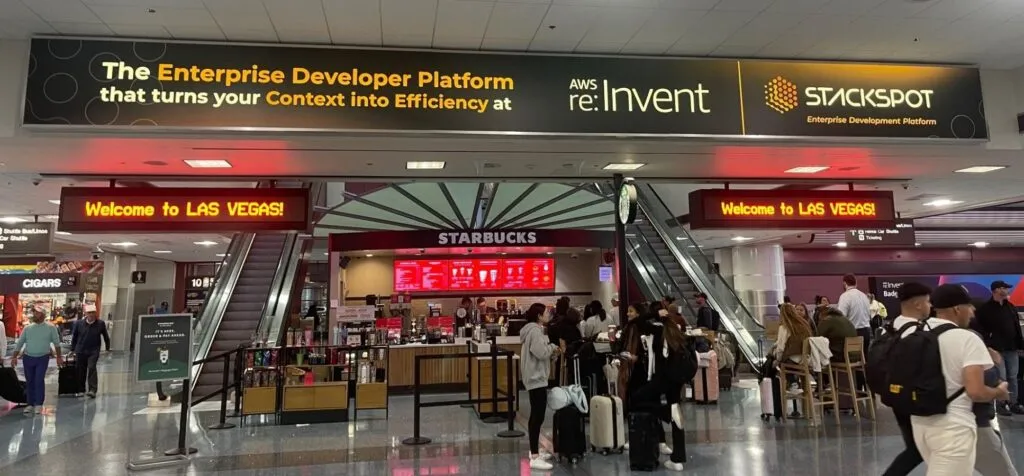 Imagem do artigo sobre AWS re:Invent 2023 com foto do saguão do aeroporto de Las Vegas, como destaque é possível ver um anúncio retangular com a frase The Enterprise Developer Platform that turns your Context into Efficiency at AWS re:Invent 2023 (este último em destaque). Ao lado da frase está a logo da empresa e StackSpot, abaixo Enterprise Development Platform. Na foto também estão diversas pessoas passando e dois painéis com Welcome to Las Vegas.