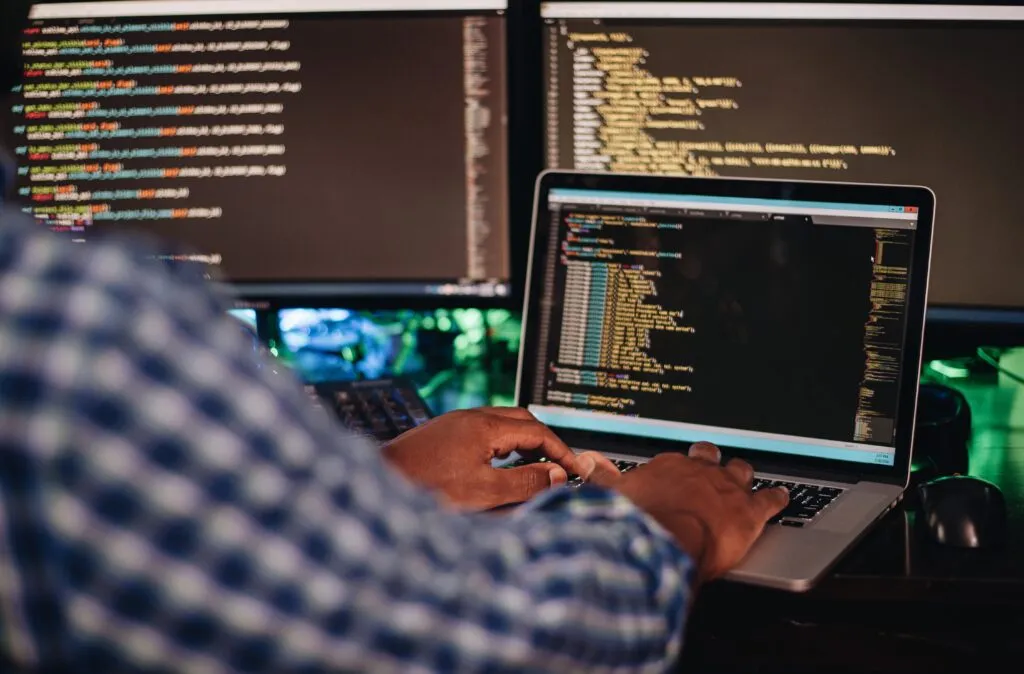 Image of a black man with his back to the wall in a blue checkered shirt, he is standing in front of three screens with yellow codes.