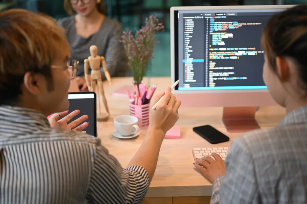 Image from the article on Gartner Top Strategic Technology Trends 2024: our HighlightsTwo Asian people in front of an office desk, sitting in their chairs, are looking at a computer screen showing computer codes. On the table are pens, a cup and saucer, a cell phone, a keyboard, another laptop, a plant and an articulated wooden doll. One of the people points a pen at the screen.