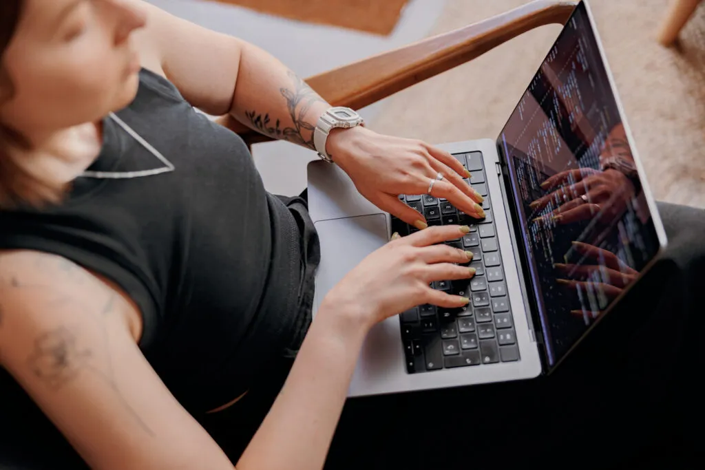 Capa do artigo "Fontes de conhecimento e o seu papel nos modelos de linguagem baseados em RAG". Na imagem, mulher branca com tatuagens nos braços e uma blusa preta está sentada com o notebook no colo. A foto foi tirada de cima, onde podemos ver as mãos da mulher no teclado e códigos na tela.