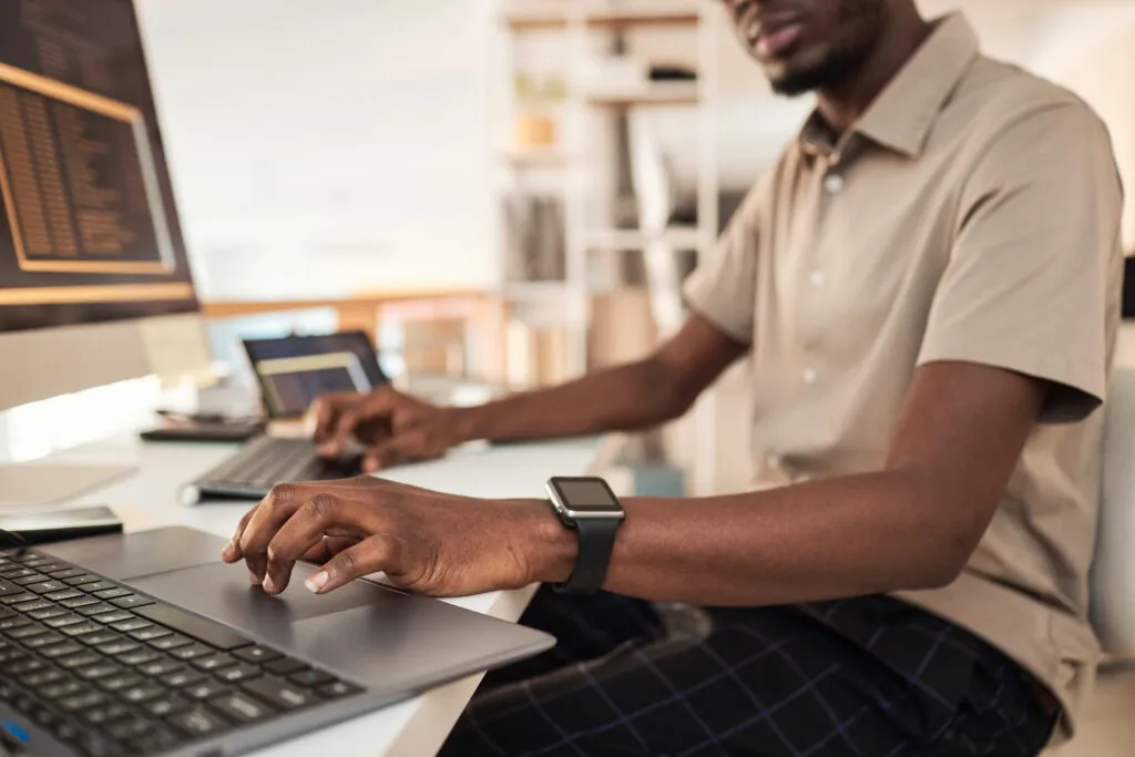 Capa do artigo Plataforma de desenvolvimento empresarial: ferramenta crucial para o sucesso da sua operação. Desenvolvedor de software, um homem negro de camisa bege, usando smartwatch trabalhando no computador.