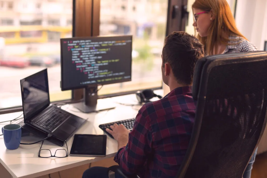 Two senior developers are working in a software development company office. Focus on the man sitting in front of two screens full of codes.