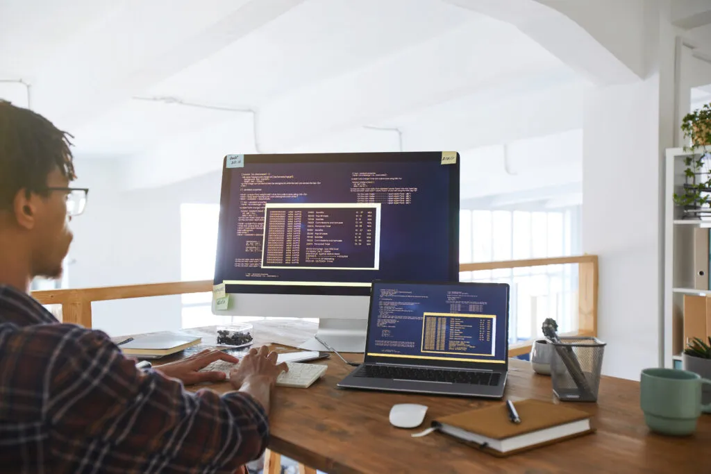 Imagem capa do conteúdo sobre grandes modelos de linguagem ou Large Language Models (LLMs), onde há uma foto de um homem negro em frente ao computador, suas mãos estão no teclado e ele observa uma tela e um notebook que mostram códigos.