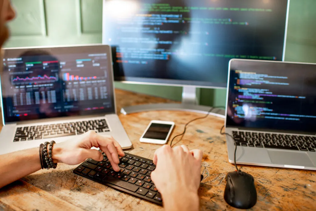 Cover of the article "Quality and efficiency: how to run automated Rest API testing with an AI tool". In the picture, young programmer writing a program code sitting at the workplace with three monitors in the office.