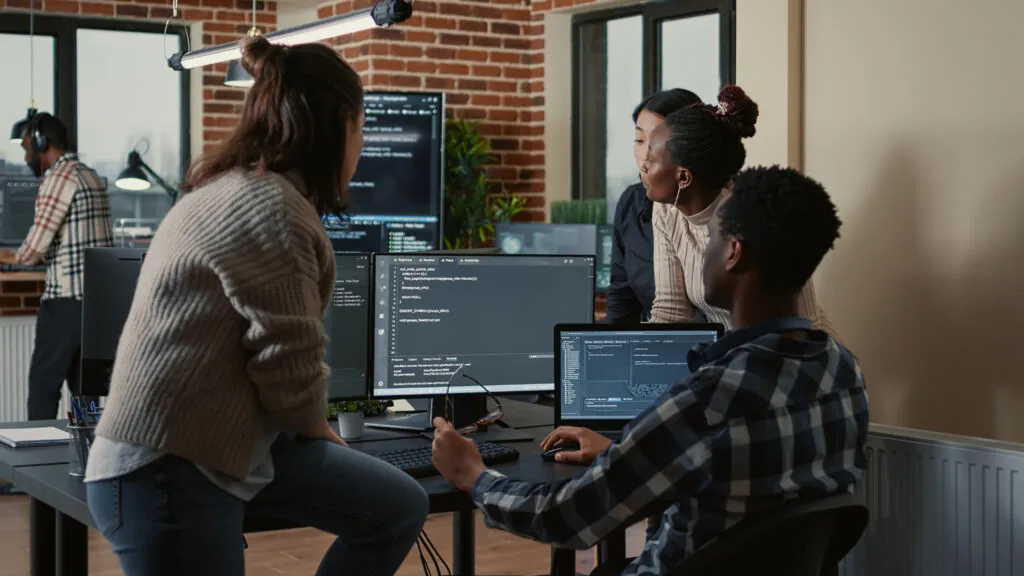 Capa do artigo "Time de engenharia de plataforma: tudo o que você precisa saber". Nela, temos quatro pessoas ao redor de uma mesa com computadores, onde o monitor mostra códigos de programação.