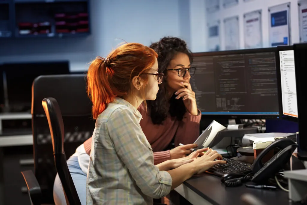 The cover of the article "Smart backlog refinement: How an AI Tool Enhances Product Management" features two white women wearing glasses, one with red hair in a ponytail and the other with loose, curly brown hair. Both are working in front of a computer monitor. Behind the brunette woman is another monitor.