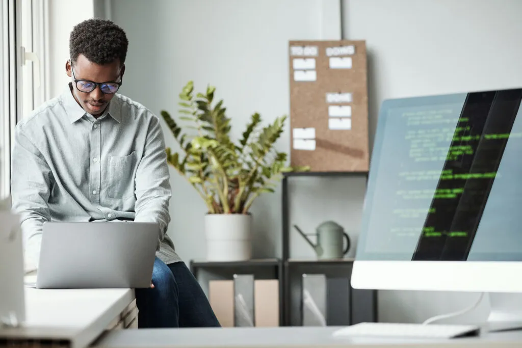 Cover of the article Contextualized AI for learning: How to transform new technical skills. The image shows a black man with short hair and glasses, wearing a light-coloured shirt and holding a laptop on a table. The environment is an office and a monitor with codes appears in the foreground.