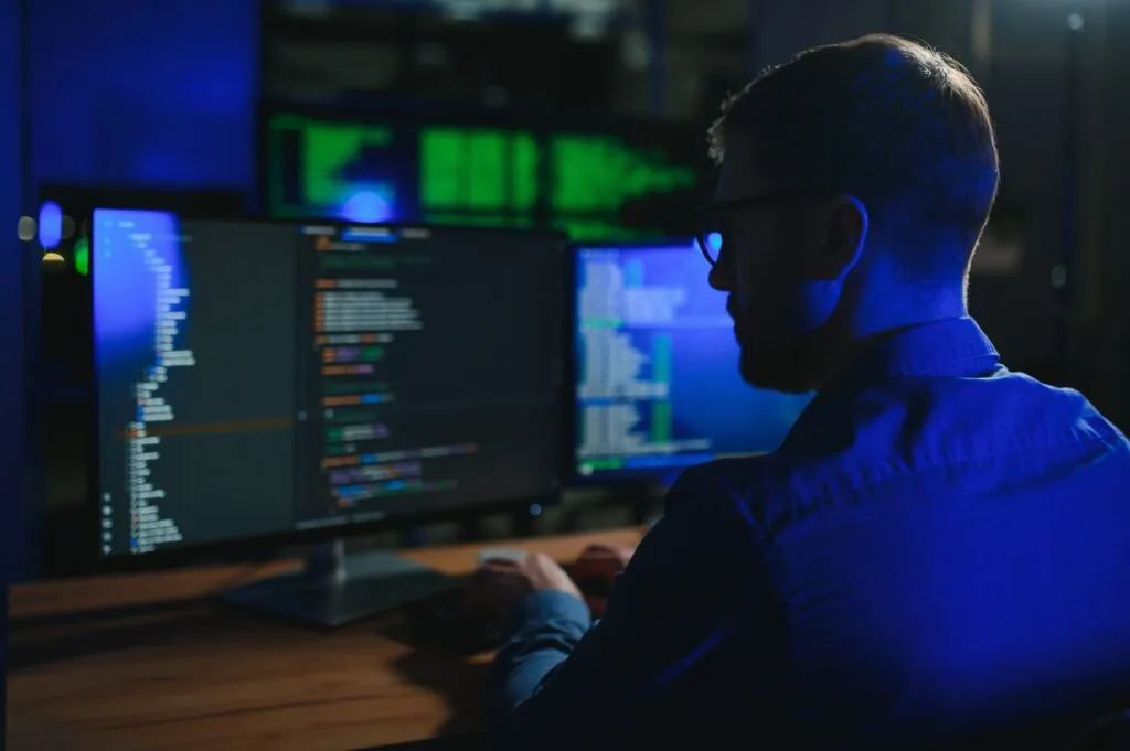 Cover of the article on AI in Technical Writing. The image shows a short-haired man wearing glasses in front of a monitor. The image is dark, with full focus on the monitor with codes.