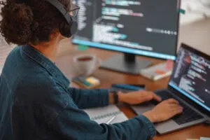 Capa do artigo sobre Testes unitários para AWS Glue Jobs. Na imagem, temos uma mulher branca usando fones, óculos e uma camisa verde de manga comprida, ela está em frente ao notebook e a um monitor que exibe códigos.