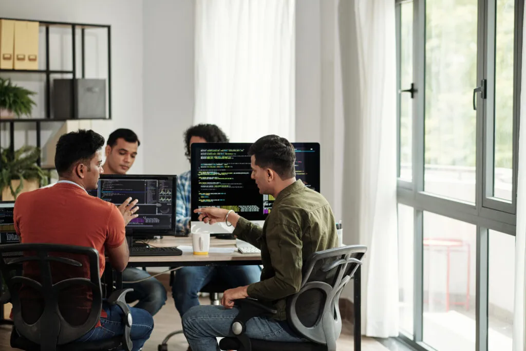 Cover of the article Code migration with AI using Fortify. The image shows an office with desks and computer monitors with developers talking. On the monitors are programming codes.