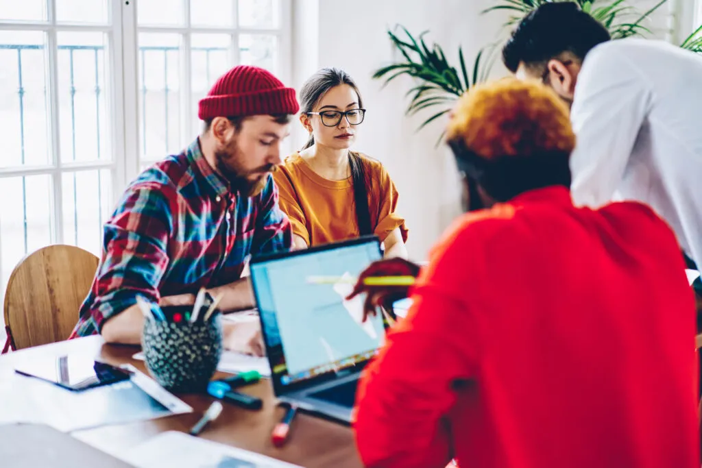 Cover of the article on AI Agents vs AI Assistants. In the image, we have a group of multi-ethnic young people in an office environment, all at a table with notebooks and office supplies.