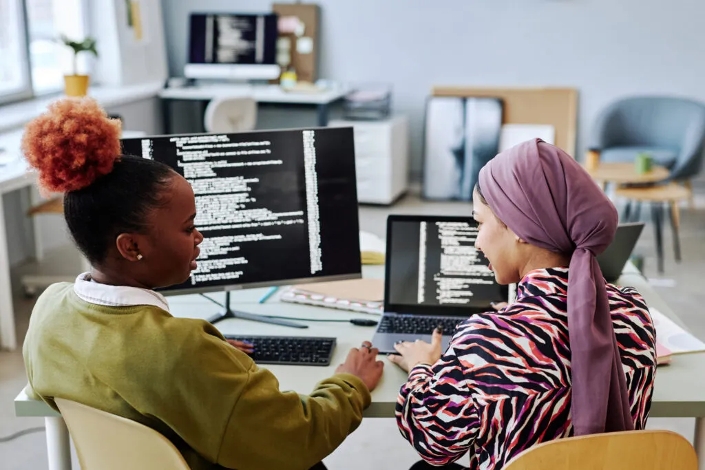 Capa do artigo "Estratégias de modernização do legado". Na imagem, temos duas mulheres, uma mulher negra de cabelos crespos e uma mulher de hijab, ambas estão em frente à mesa de trabalho com dois monitores que exibem códigos.