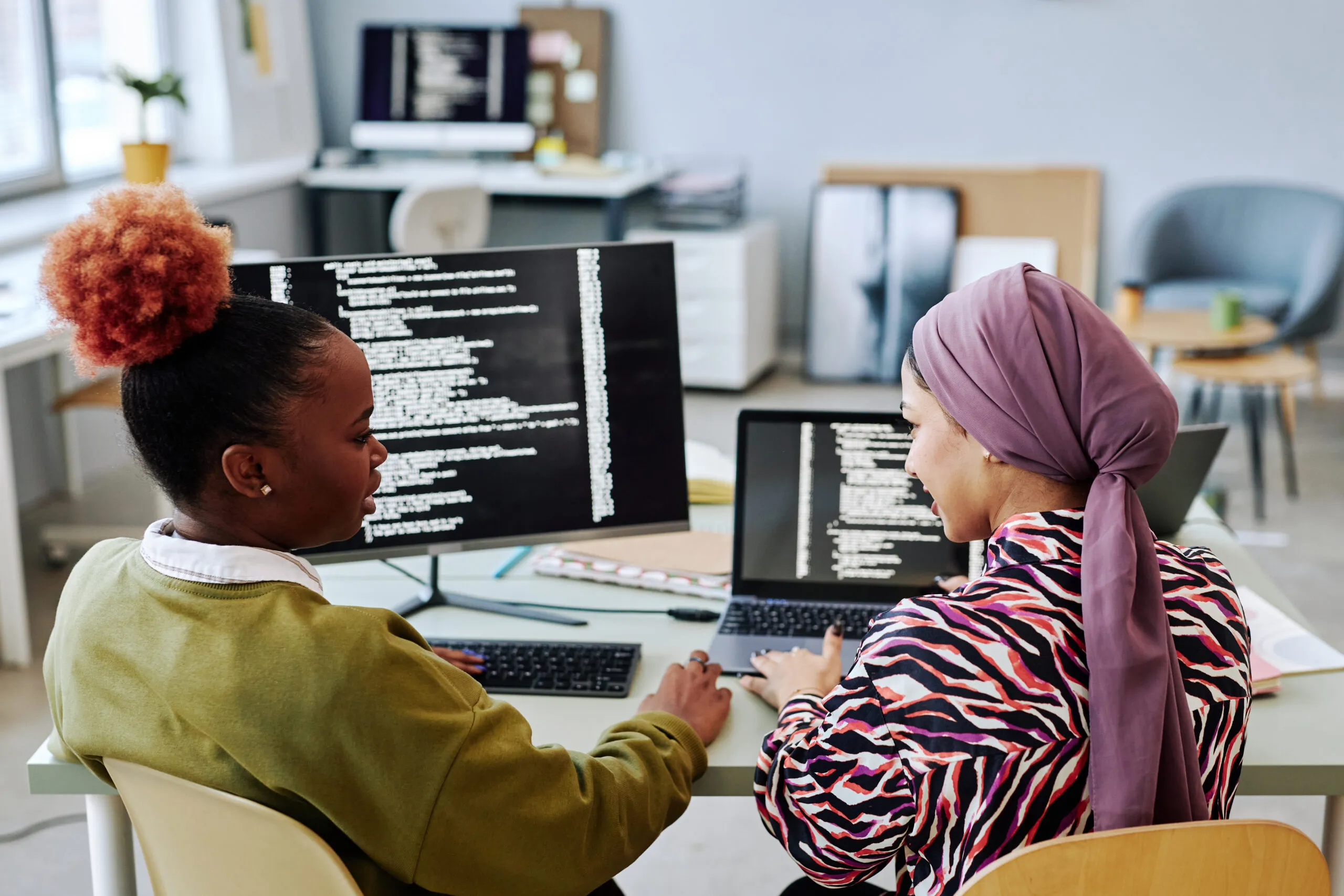 Capa do artigo "Estratégias de modernização do legado". Na imagem, temos duas mulheres, uma mulher negra de cabelos crespos e uma mulher de hijab, ambas estão em frente à mesa de trabalho com dois monitores que exibem códigos.