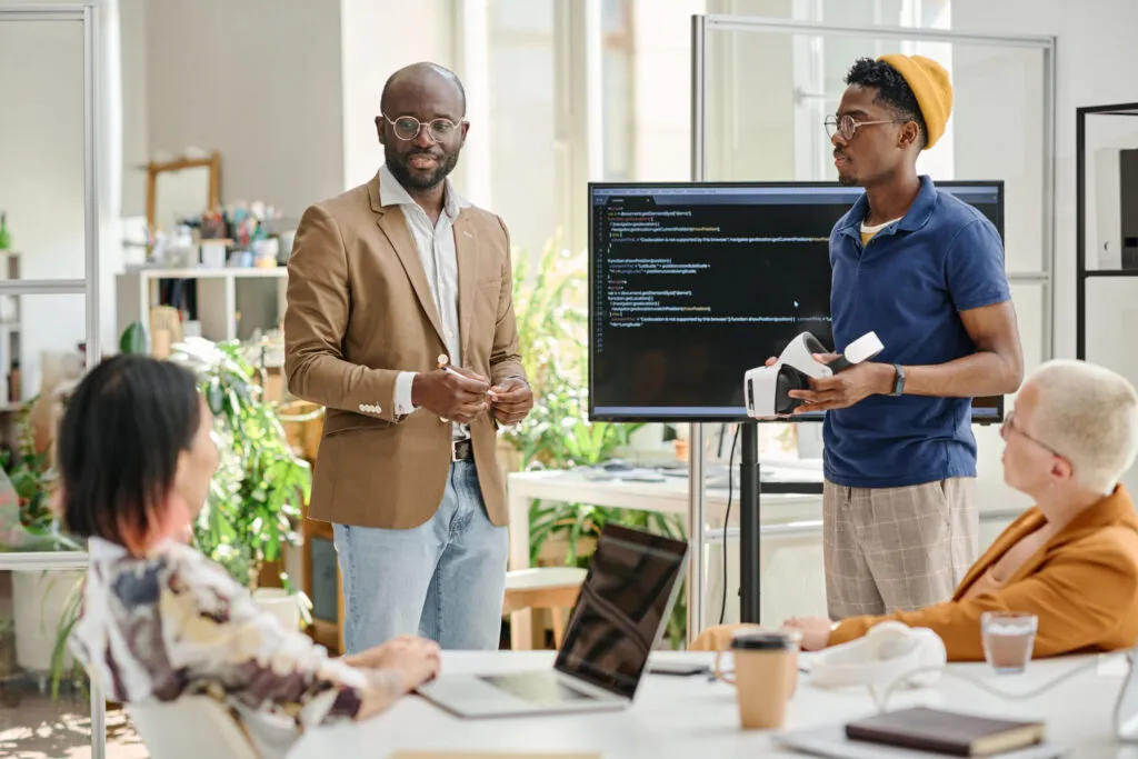 Cover of the backlog refinement case article. The image shows a development team in an office environment. Two people are standing and two are sitting. The standing ones seem to be presenting something, as there is a monitor behind them.