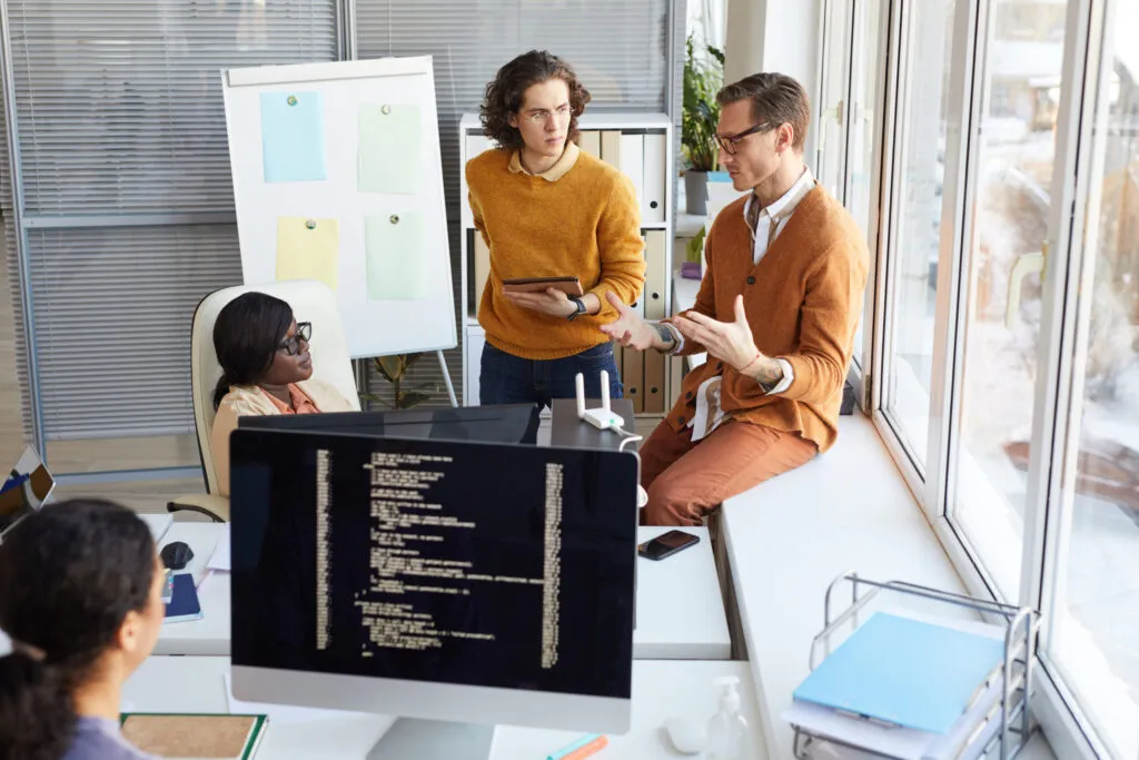 Cover of TMMi article with AI. It shows two young white men standing, while a black woman is sitting. They are in an office and appear to be talking.