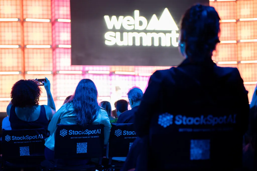 A scene from Web Summit Rio 2025 captured during one of the main sessions. The stage screen in the background displays “Web Summit,” while in the foreground, a woman wearing a StackSpot AI cape sits facing the stage, her back to the camera.