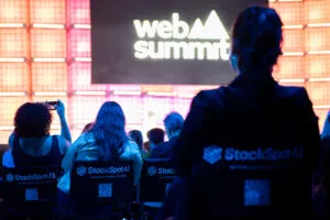 A scene from Web Summit Rio 2025 captured during one of the main sessions. The stage screen in the background displays “Web Summit,” while in the foreground, a woman wearing a StackSpot AI cape sits facing the stage, her back to the camera.