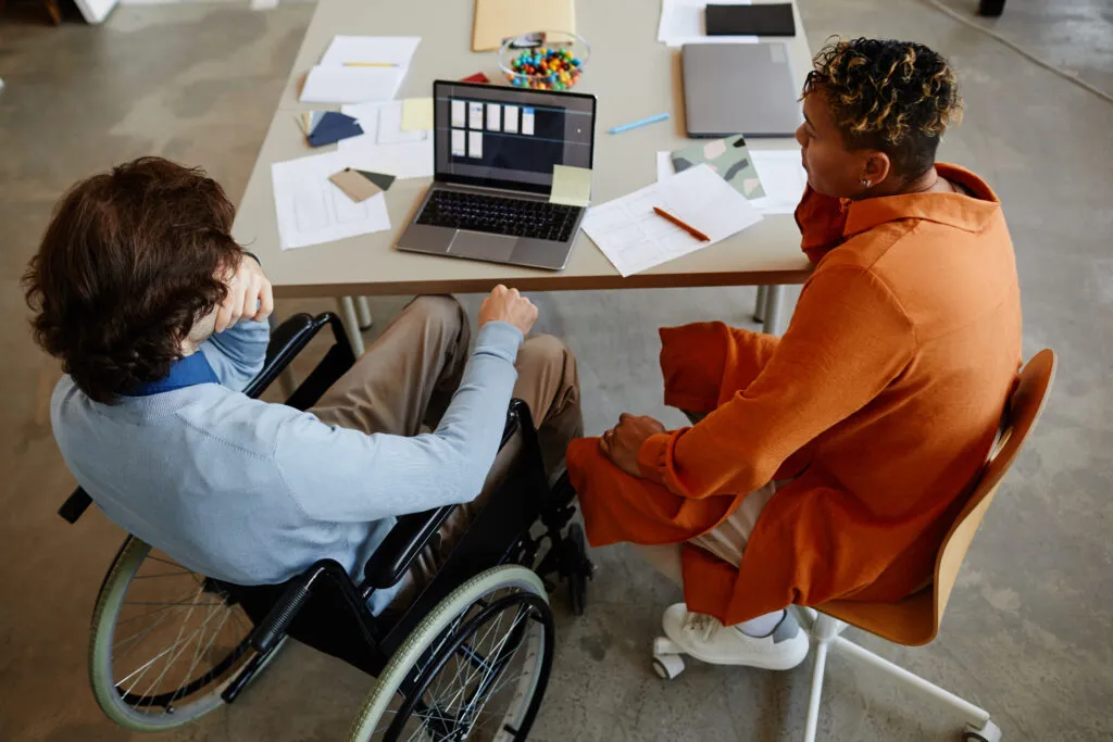 Capa do artigo sobre modelos LLMs. Na imagem, temos duas pessoas em um ambiente de escritório. Uma pessoa está em uma cadeira de rodas em frente a um notebook e a outra pessoa está ao seu lado usando uma roupa laranja chamativa.
