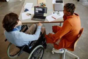 Capa do artigo sobre modelos LLMs. Na imagem, temos duas pessoas em um ambiente de escritório. Uma pessoa está em uma cadeira de rodas em frente a um notebook e a outra pessoa está ao seu lado usando uma roupa laranja chamativa.
